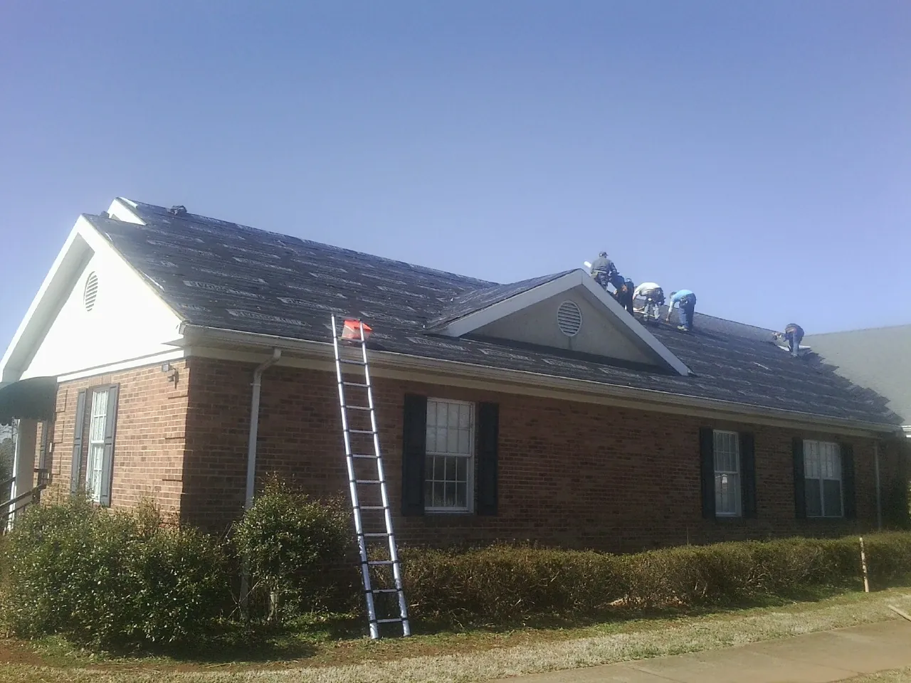 Crew tearing shingles off a brick ranch roof with a ladder set — Metal Roof Repair work by our crew in Bay City